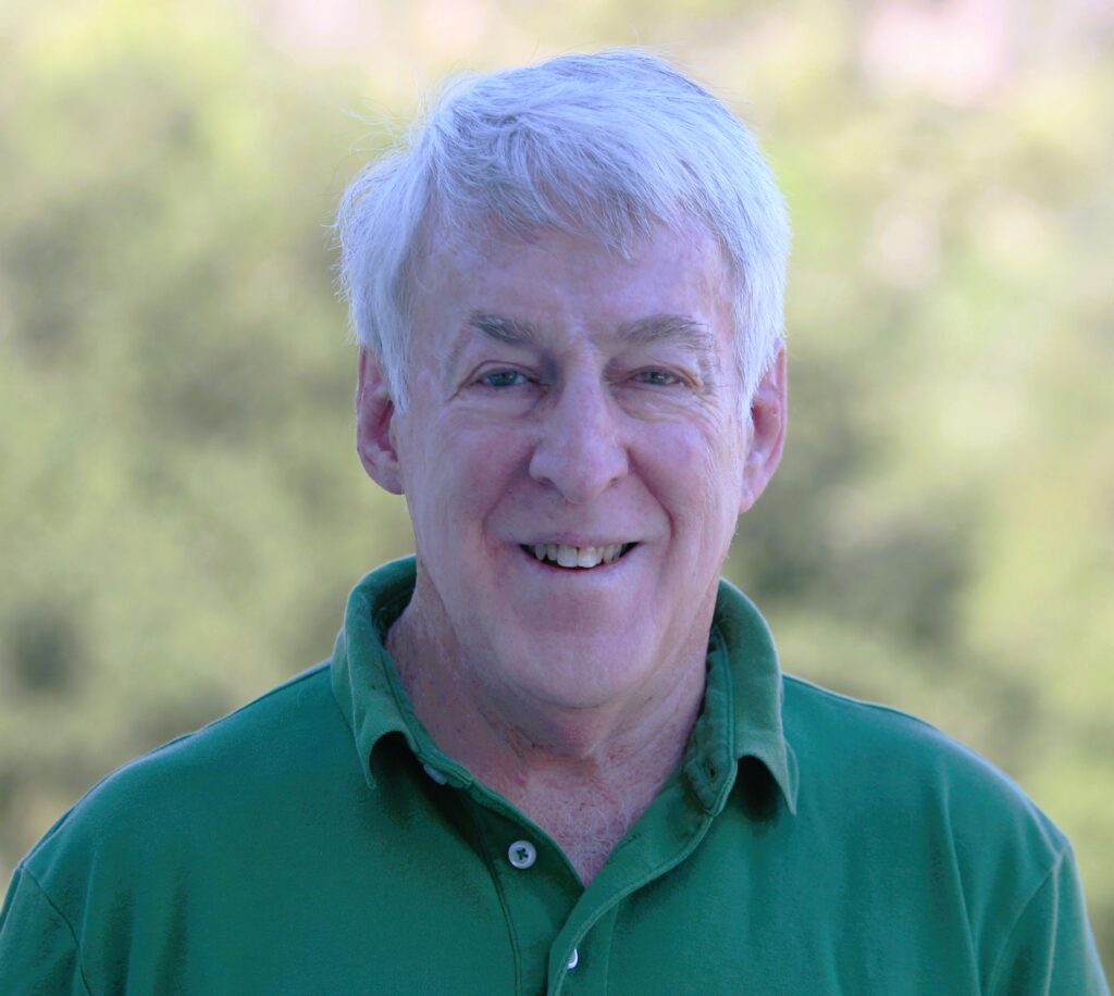 Mike Simmons is wearing a green polo shirt and smiling in in this head and shoulders photo. There is plant and tree foliage in the background.