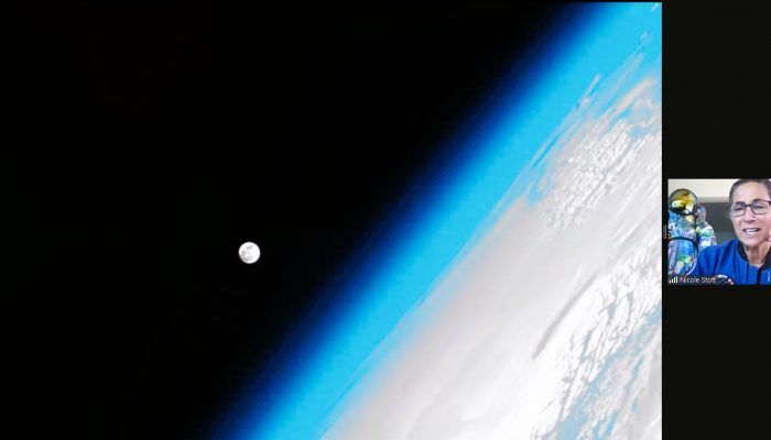 Astronomy for Equity. NASA Astronaut Nicole Stott gives an online live presentation in Libya, Africa. The screen shows a huge picture of Earth from space, off to the side, with Earth's atmosphere glowing in bright light blue along the border of the planet. The moon is a large dot against the blackness of space and next to Earth.