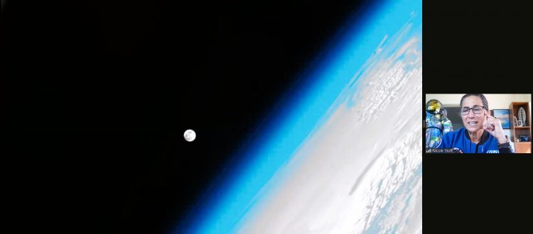 Astronomy for Equity. NASA Astronaut Nicole Stott gives an online live presentation in Libya, Africa. The screen shows a huge picture of Earth from space, off to the side, with Earth's atmosphere glowing in bright light blue along the border of the planet. The moon is a large dot against the blackness of space and next to Earth.