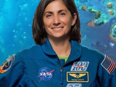 Astronomy for Equity. Former NASA Astronaut Nicole Stott is smiling and wearing a blue jumpsuit that has many patches on it including the American flag and NASA patches. She is standing in front of a blue-green background.