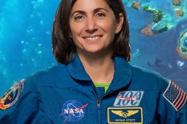 Astronomy for Equity. Former NASA Astronaut Nicole Stott is smiling and wearing a blue jumpsuit that has many patches on it including the American flag and NASA patches. She is standing in front of a blue-green background.