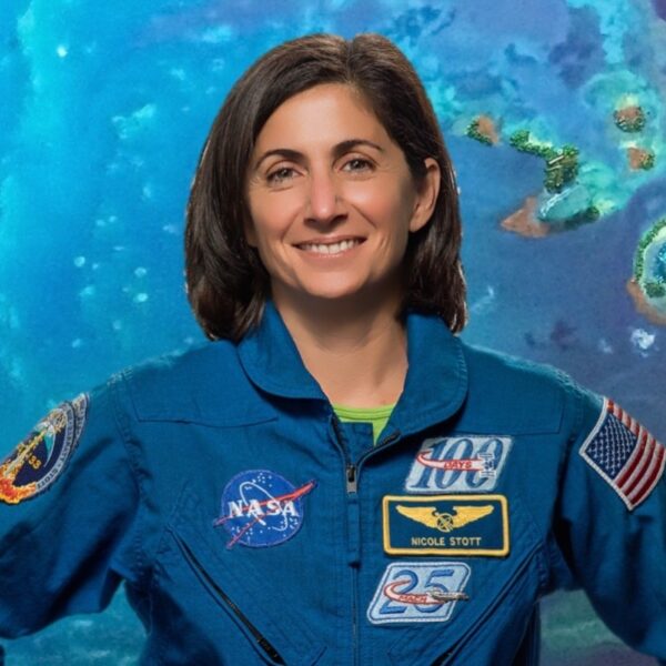 Astronomy for Equity. Former NASA Astronaut Nicole Stott is smiling and wearing a blue jumpsuit that has many patches on it including the American flag and NASA patches. She is standing in front of a blue-green background.