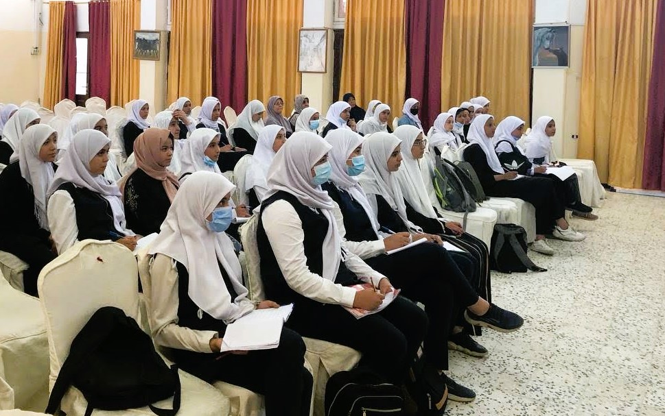 About fifty female students in Libya sit in a small auditorium as they watch a presentation about astronomy. The girls are dressed in matching clothes and head coverings.