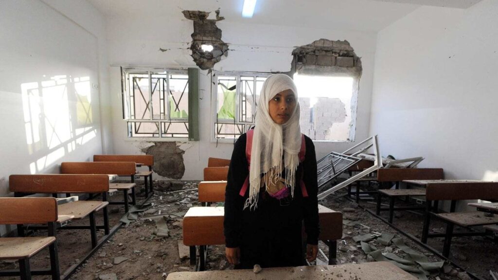 A young woman in Libya stands in an empty classroom that was devastated by civil warfare. There is much rubble in the room. She looks sad.