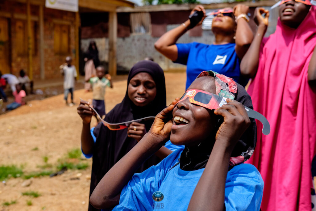 An African child and two African teens try on paper eclipse glasses while another African child tries to figure hers out. They are standing in dirt at a camp for internally displaced persons.