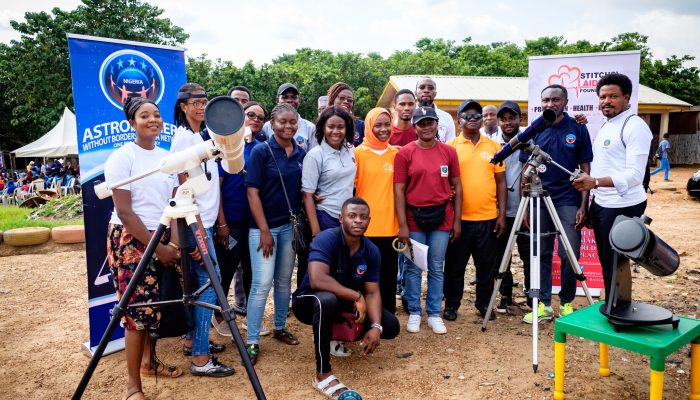 Seventeen dark-skinned adults smile for a photo at a dirt camp with tents, signs, and some grass. There are three telescopes.