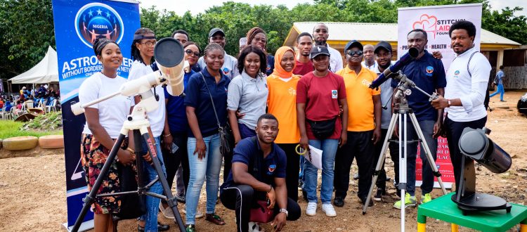 Seventeen dark-skinned adults smile for a photo at a dirt camp with tents, signs, and some grass. There are three telescopes.
