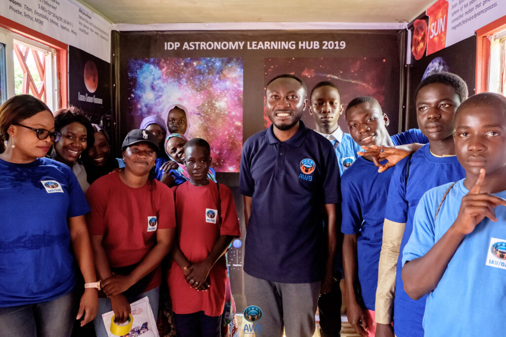 The Learning Hub team is shown inside the Learning Hub with some of the IDP children. There are posters of astronomical objects on the walls behind them.