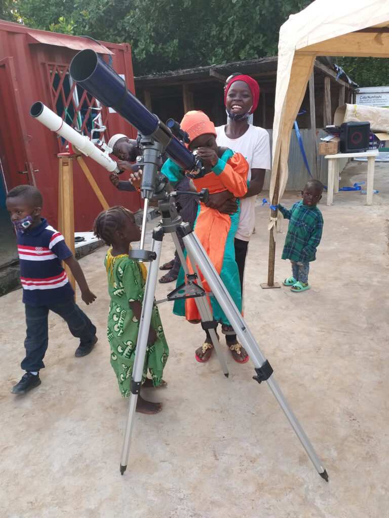 Outside the Learning Hub, a mother lifts her young child to the eyepiece of a telescope to view the Moon. There is another telescope in the background and other children waiting. 