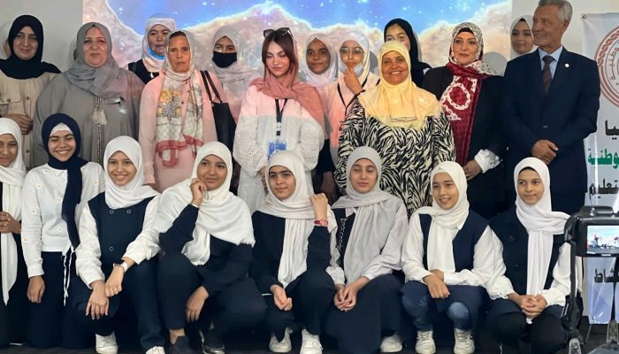 Twenty-three Libyan women and girls with head coverings and a Libyan man in a suit pose for a photo. Behind them is a backdrop with a nebula and galaxies.