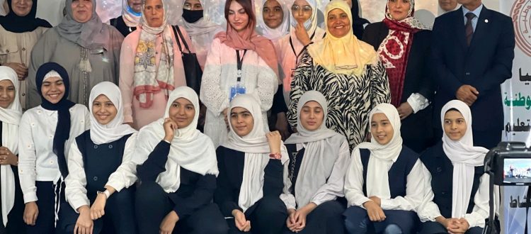 Twenty-three Libyan women and girls with head coverings and a Libyan man in a suit pose for a photo. Behind them is a backdrop with a nebula and galaxies.
