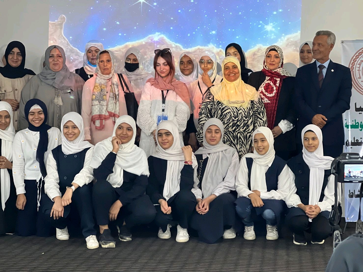 Twenty-three Libyan women and girls with head coverings and a Libyan man in a suit pose for a photo. Behind them is a backdrop with a nebula and galaxies.