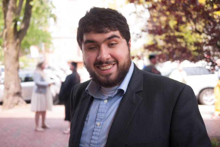 In this upper body photo, Sina Bahram looks attractive with a charming smile and a welcoming demeanor. He is wearing a light blue collar shirt and a dark blazer. He has dark hair, a beard, and one eye that closes more than the other. He is outdoors against a blurred background of people, cars, and trees.