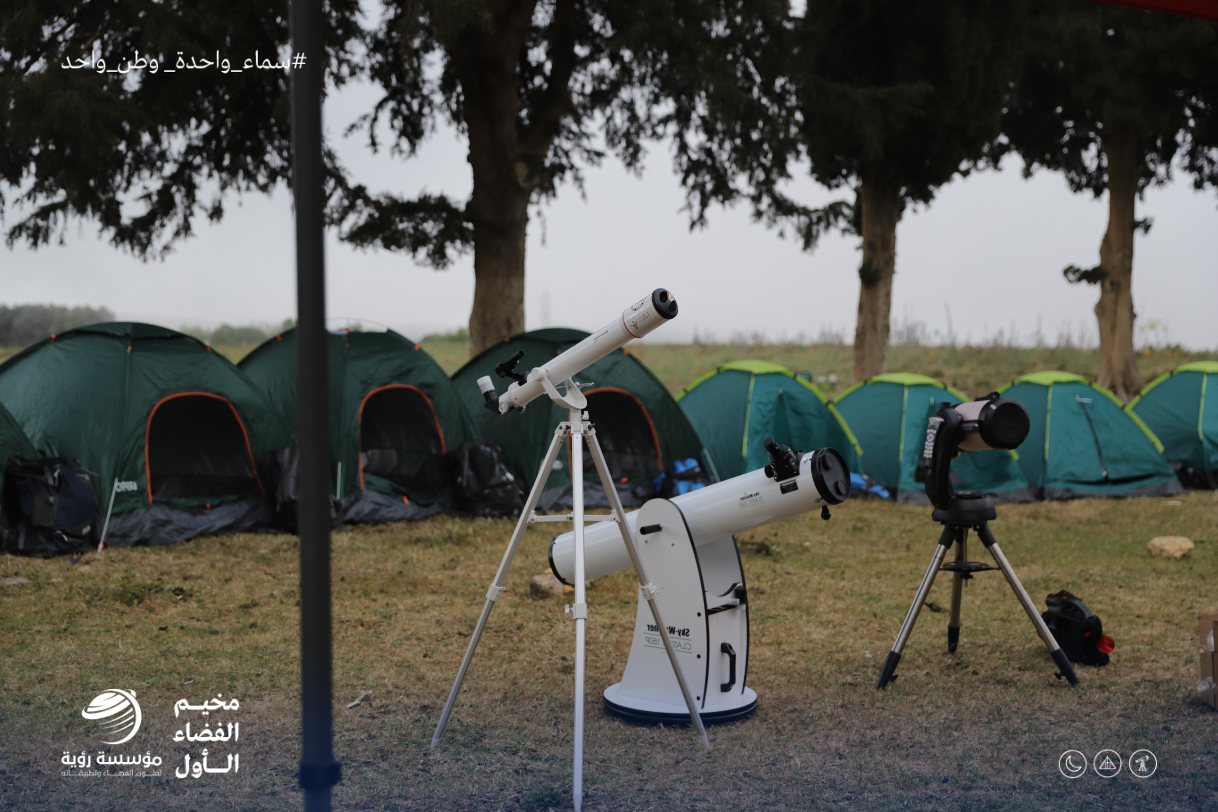 Three telescopes are lined up on a grassy field in front of a row of small tents at a space camp for students.